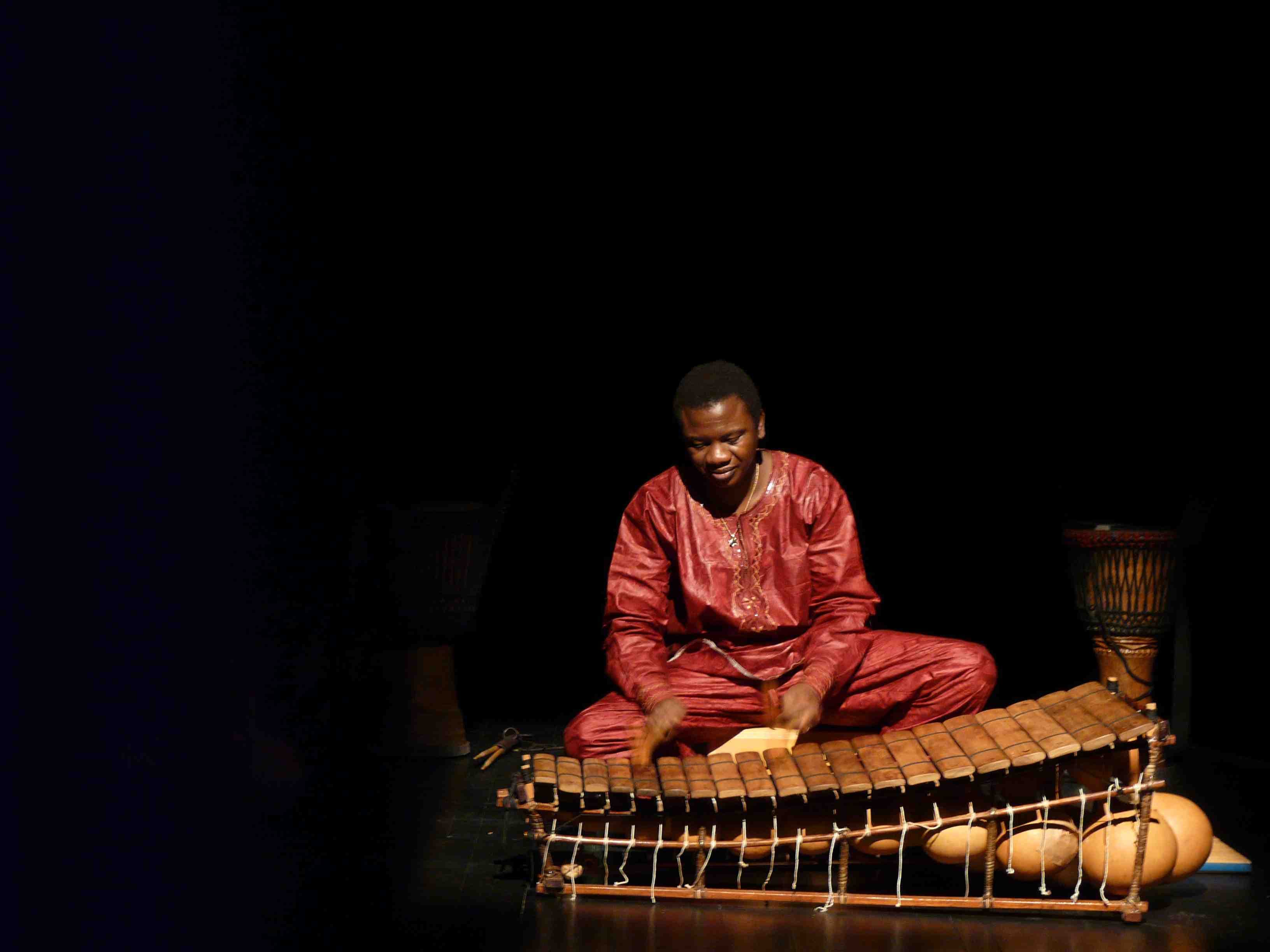 Moussa Hema jouant du balafon sur scène, en boubou rouge, entouré de djembés